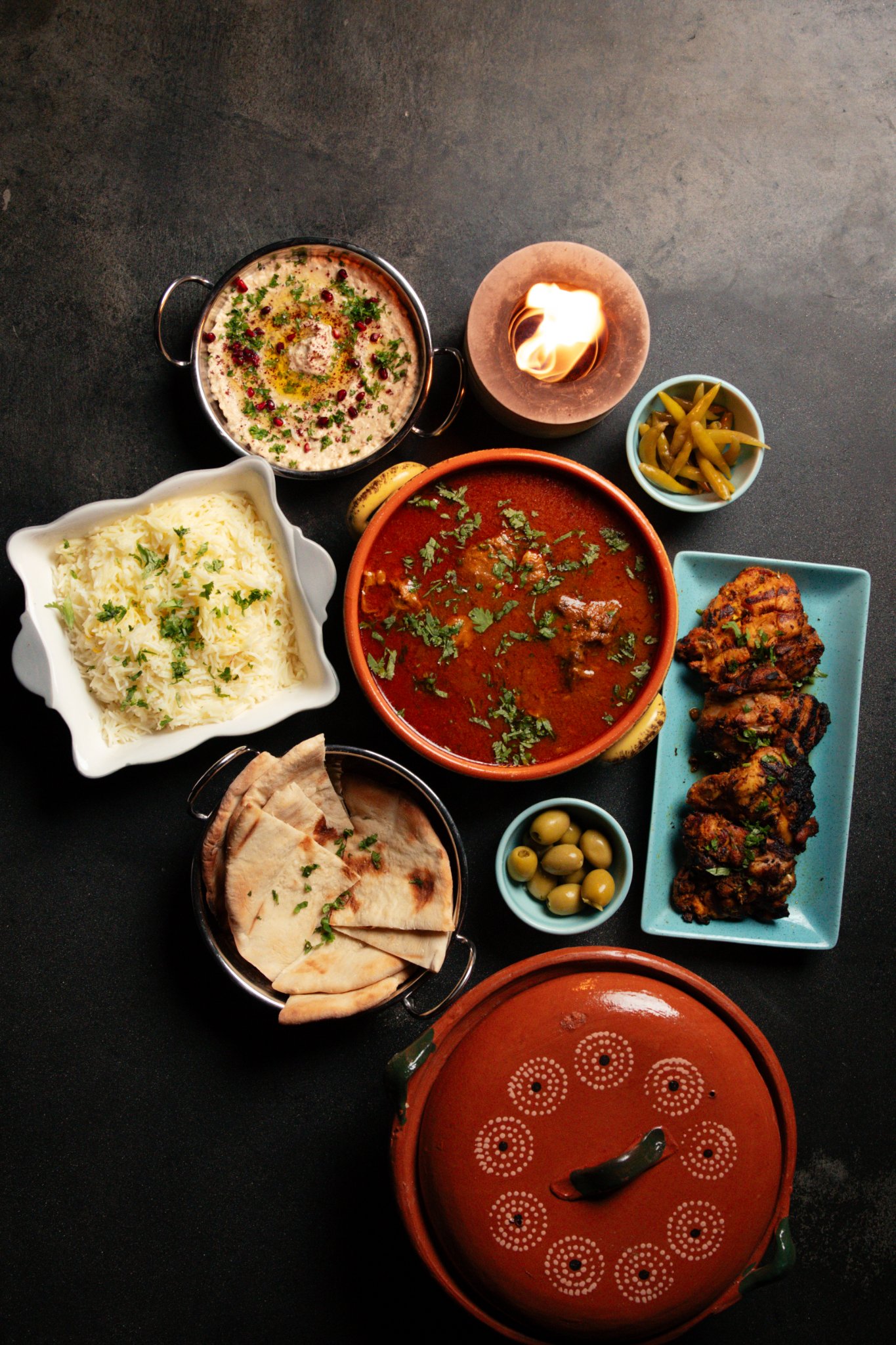 A full Goat's Haven dinner spread from above — curry, rice, grilled chicken, warm flatbread, dips, olives and pickles around a small flame