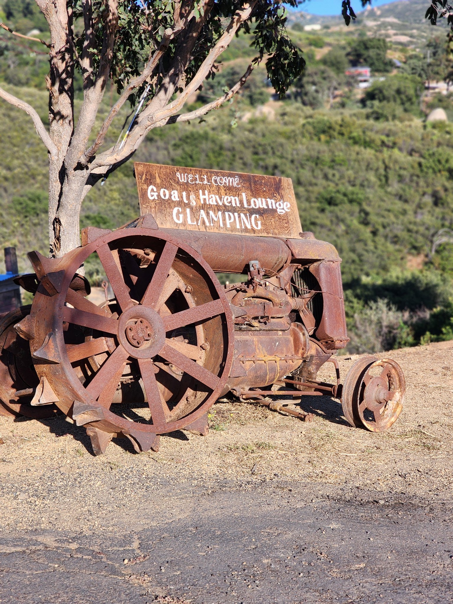 The wooden &lsquo;Welcome — Goat&rsquo;s Haven Lounge Glamping&rsquo; sign on the old rusted tractor at the entrance, hills behind